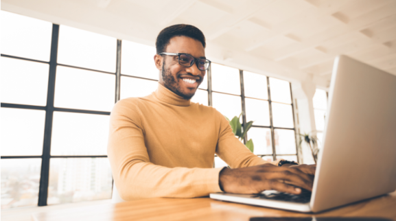 homem de óculos, sorrindo na frente de um notebook. banner post como ser corretor de imóveis